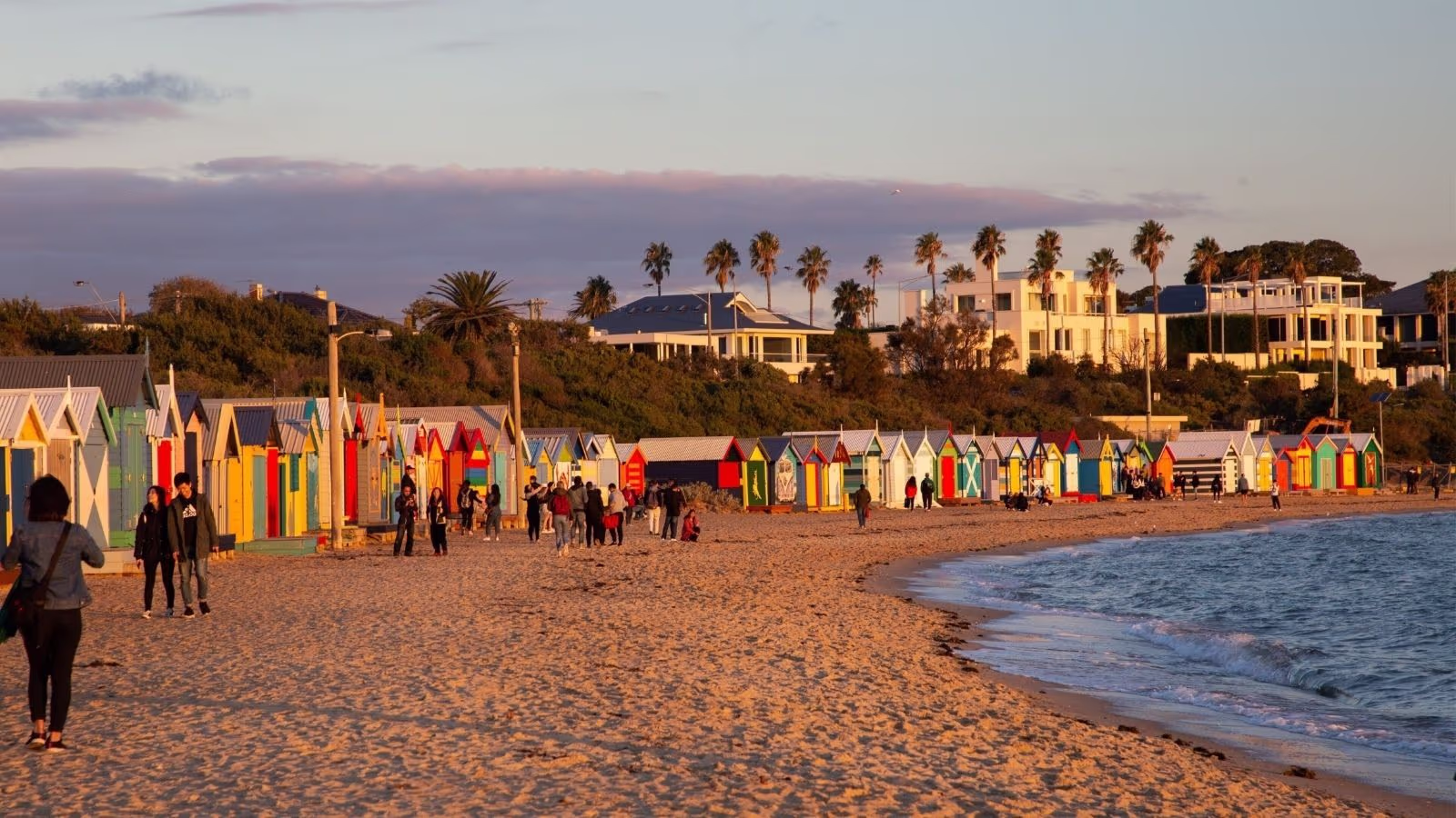 Row of brightly painted bathing boxes on the sand at Brighton Beach, Melbourne, with the city skyline behind